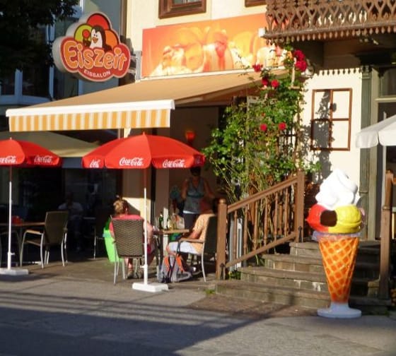 Das Bild zeigt die Gelateria Del Corso von außen. Die Fassade ist weiß, während große orangefarbene Akzente, darunter eine orange Markise, dem Gebäude einen lebendigen Look verleihen. Vor der Tür steht ein großes Modell eines Eises, und das Logo mit einem