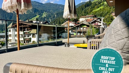 Blick auf eine Dachterrasse mit Sonnenschirmen, umgeben von Bergen und Häusern in einer Bergstadt, sonniger Tag.