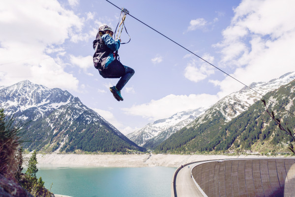 Eine Person fährt mit einer Zipline über einen türkisfarbenen Stausee. Im Hintergrund sind hohe, teils schneebedeckte Berge und ein bewaldetes Tal zu sehen. Rechts im Bild verläuft die geschwungene Staumauer entlang des Sees unter einem leicht bewölkten H