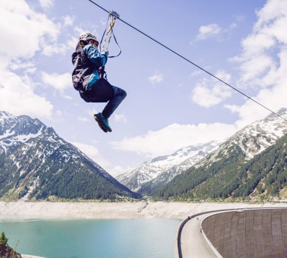 Eine Person fährt mit einer Zipline über einen türkisfarbenen Stausee. Im Hintergrund sind hohe, teils schneebedeckte Berge und ein bewaldetes Tal zu sehen. Rechts im Bild verläuft die geschwungene Staumauer entlang des Sees unter einem leicht bewölkten H