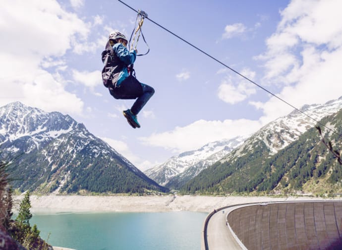 A person is riding a zipline high above a turquoise reservoir. In the background, tall, partly snow-covered mountains and forested slopes frame the valley. On the right side of the image, a curved dam runs along the lake under a lightly clouded sky.