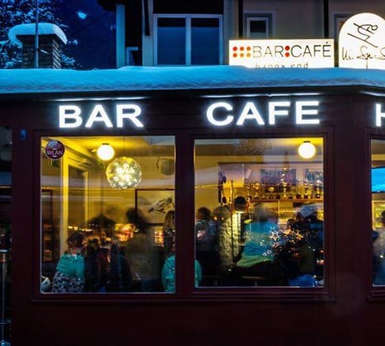 Das Bild zeigt das Happy End bei Nacht. Das rote Gebäude ist von Schnee umgeben, und durch die großen Fenster kann man sehen, wie Menschen drinnen gemütlich sitzen. Die warme Beleuchtung im Inneren bildet einen schönen Kontrast zur dunklen Winterumgebung.