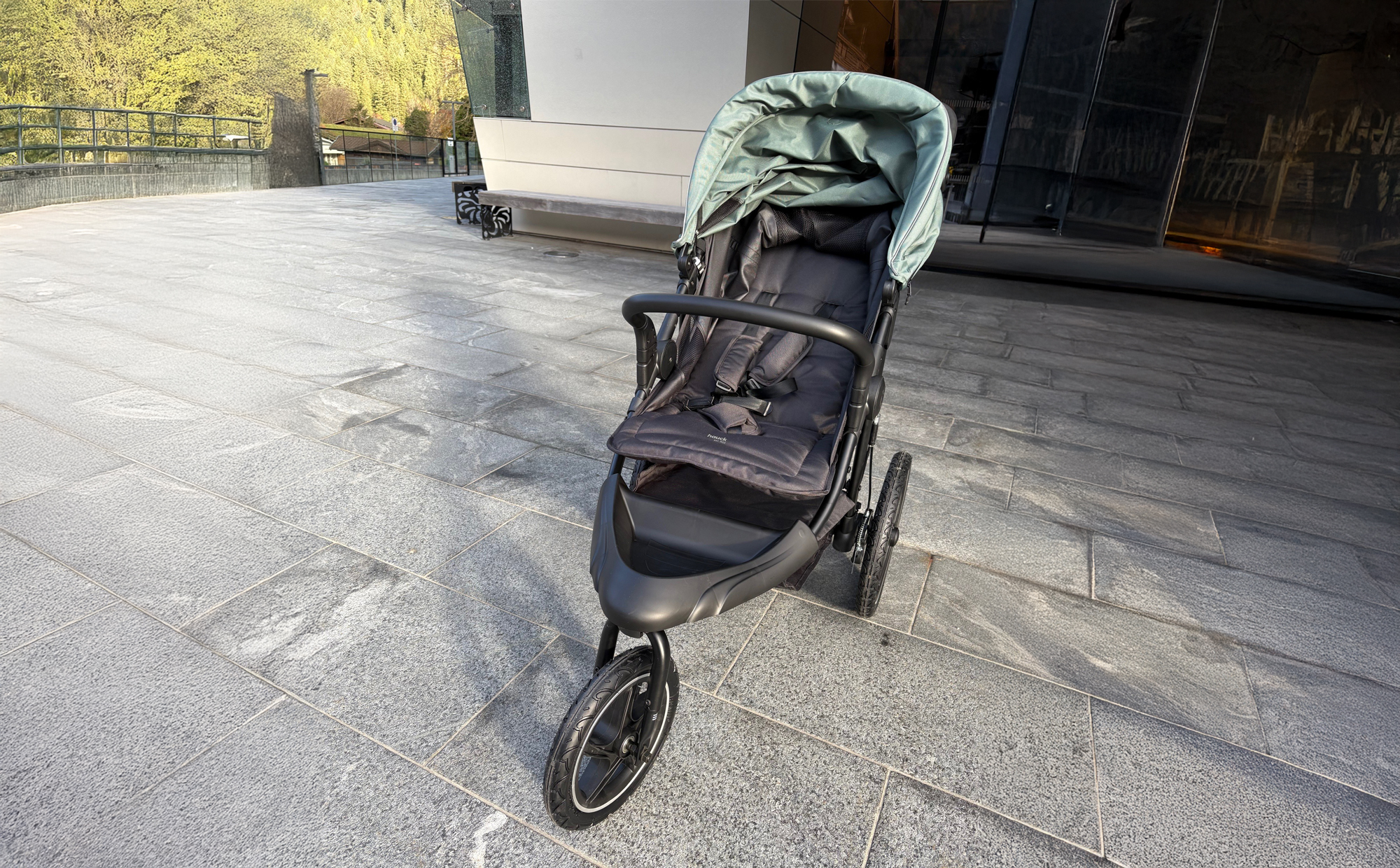 Ein schwarzer Kinderwagen mit grünem Verdeck steht auf einem breiten, glatten Gehweg vor einem modernen Gebäude in Mayrhofen. Der Zugang ist ebenerdig und gut befahrbar.