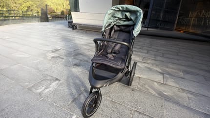 Ein schwarzer Kinderwagen mit grünem Verdeck steht auf einem breiten, glatten Gehweg vor einem modernen Gebäude in Mayrhofen. Der Zugang ist ebenerdig und gut befahrbar.