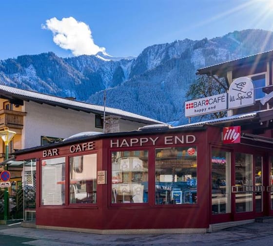 Das Bild zeigt das Happy End von außen – ein auffälliges rotes Gebäude. Dahinter stehen zwei weitere Häuser. Im Hintergrund erheben sich Berge, umgeben von Bäumen. Der Himmel ist blau mit einer einzelnen weißen Wolke.
