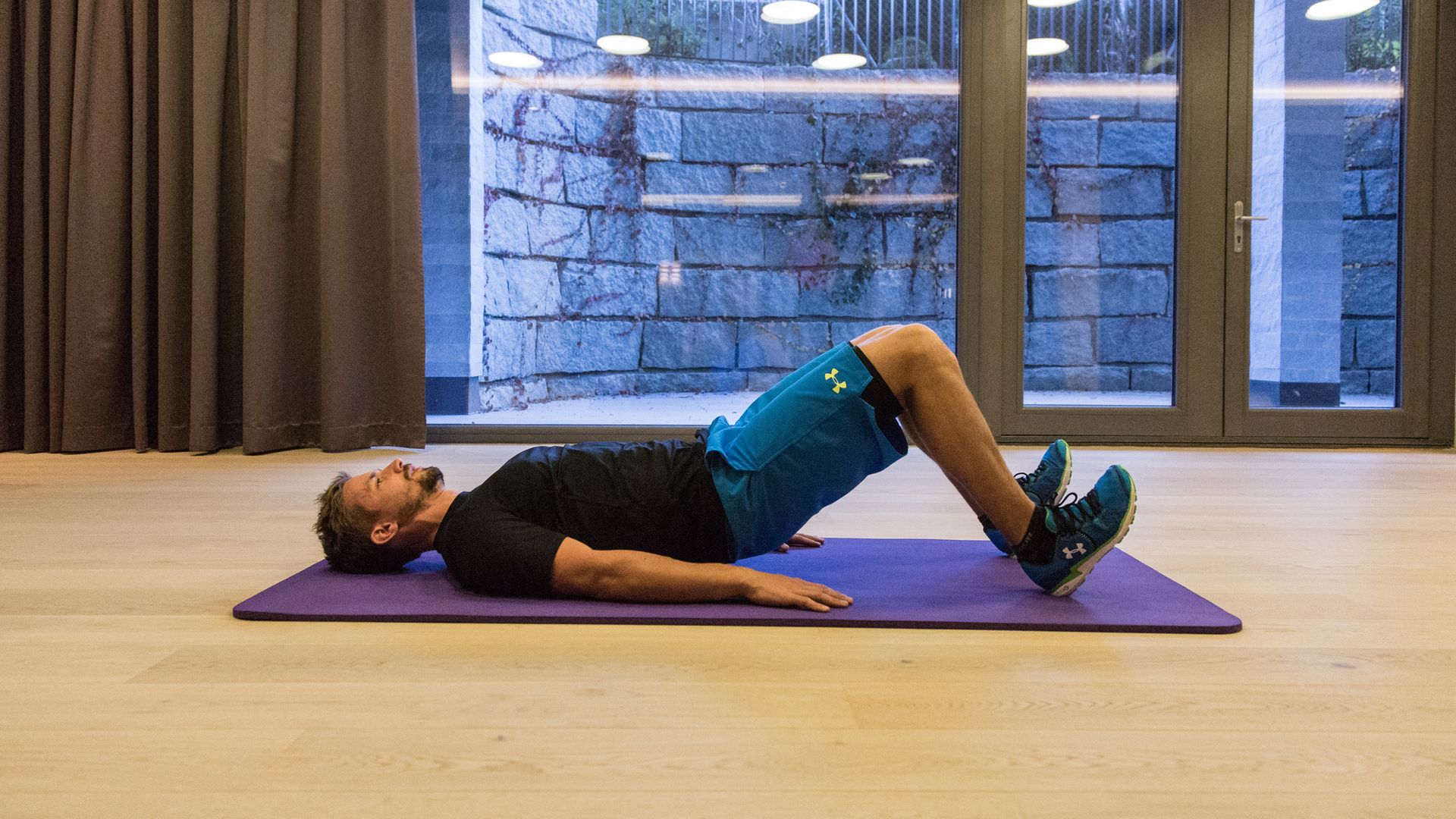 Mann führt Hüftbrücke auf Yogamatte in Trainingshalle in Mayrhofen aus. Ski-Vorbereitungstraining mit Christoph Ebenbichler – stärkt Rücken, Beine und Rumpf für einen fitten Start in den Winter.