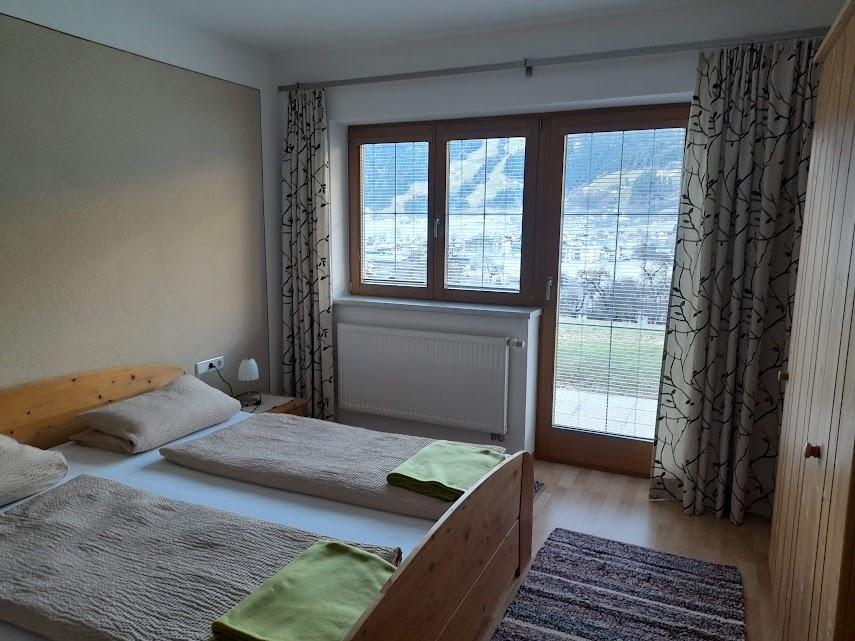 Bedroom with two beds, large windows, curtains, balcony, wooden furniture, and a rug.