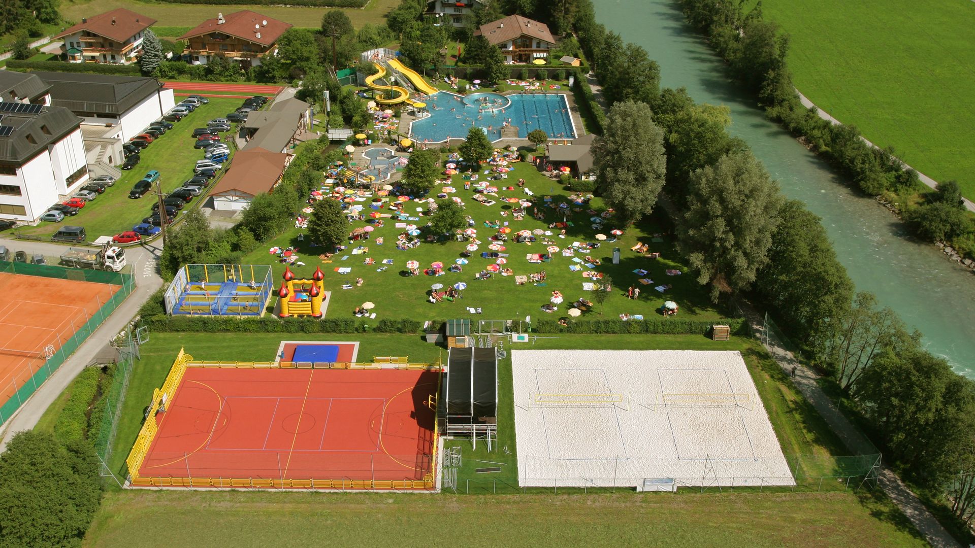 Aerial view of the Fun Court at Sommerwelt Hippach with tennis court, hard court with basketball hoops and goals, beach volleyball facility, trampolines, climbing tower, sand play area, and outdoor pool – surrounded by green landscape along the riverside.