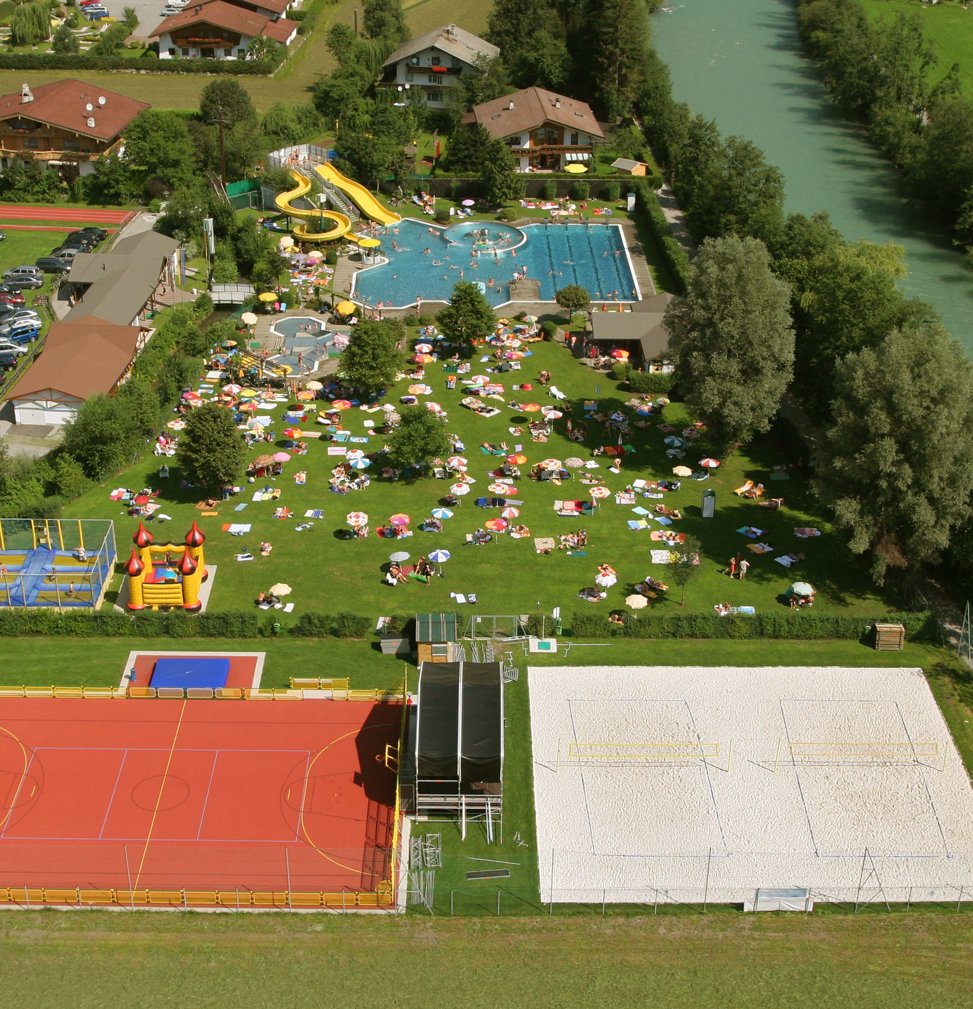 Luftaufnahme des Fun-Courts in der Sommerwelt Hippach mit Tennisplatz, Hartgummiplatz mit Basketballkörben und Toren, Beachvolleyball-Anlage, Trampolinen, Kletterturm, Sandwelt und Freibad – eingebettet in grüne Landschaft am Flussufer.
