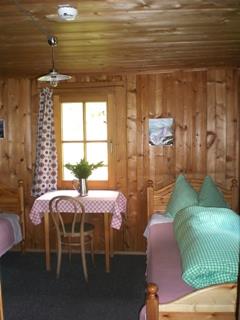 Cozy wooden room with window, table with red checkered cloth, chair, plant, and couch with green pillows.