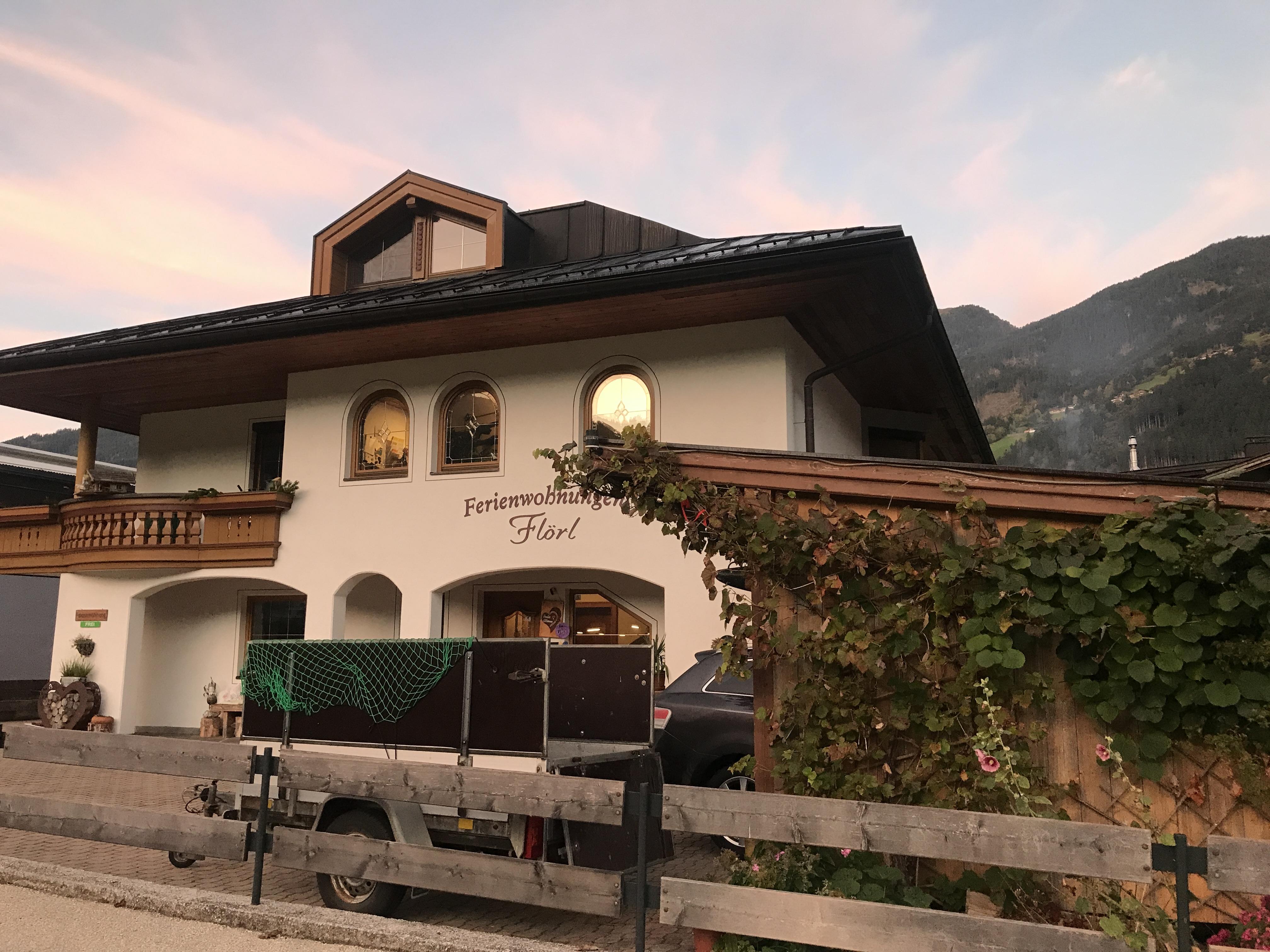 Häuser mit traditioneller Architektur vor Bergkulisse bei Sonnenuntergang, mit Pflanzen und Fahrzeugen im Vordergrund.