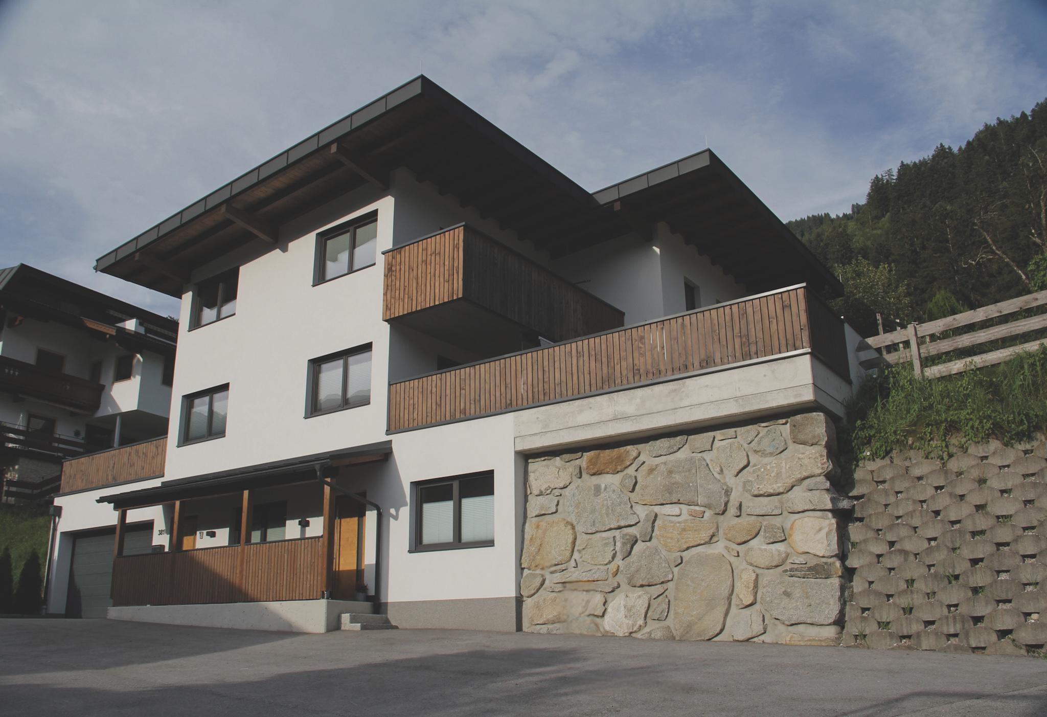 Modernes Mehrfamilienhaus mit weißen Wänden, Holzverkleidungen und steiniger Sockel, vor Himmel und Wald.