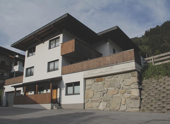 Modernes Mehrfamilienhaus mit weißen Wänden, Holzverkleidungen und steiniger Sockel, vor Himmel und Wald.