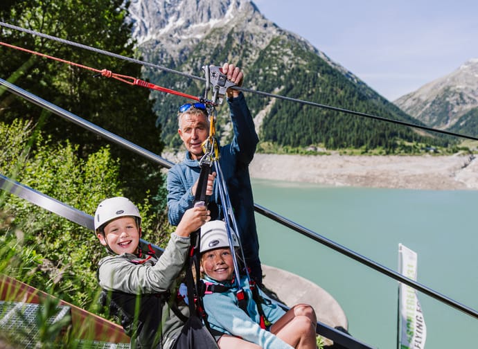 A man and two children with helmets sit on a metal platform near mountains, ready for the Schlegeis 131 Flying Fox.