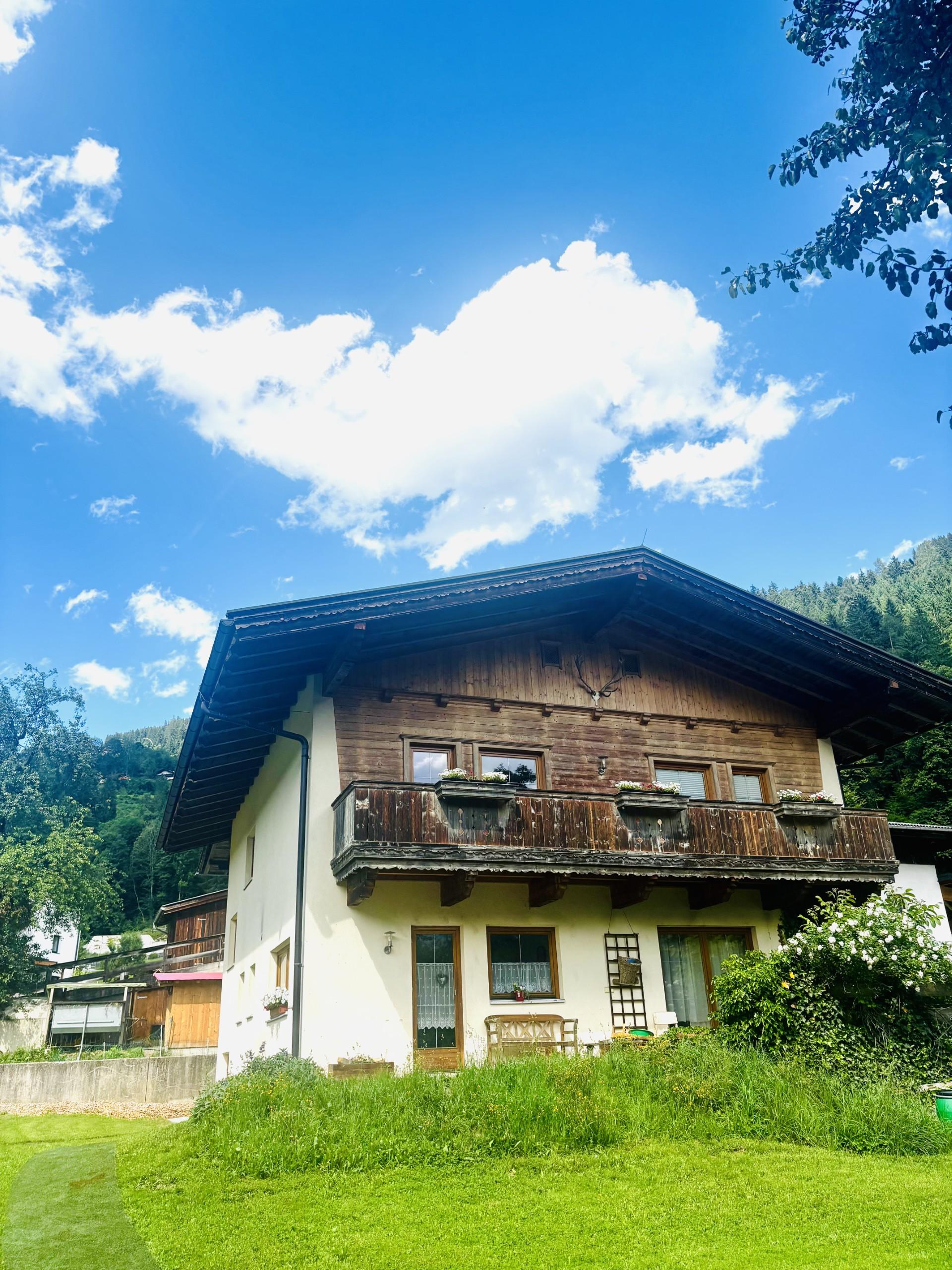 Ein zweistöckiges Haus mit Holzfassade, Balkon und Garten vor einem blauen Himmel mit Wolken.