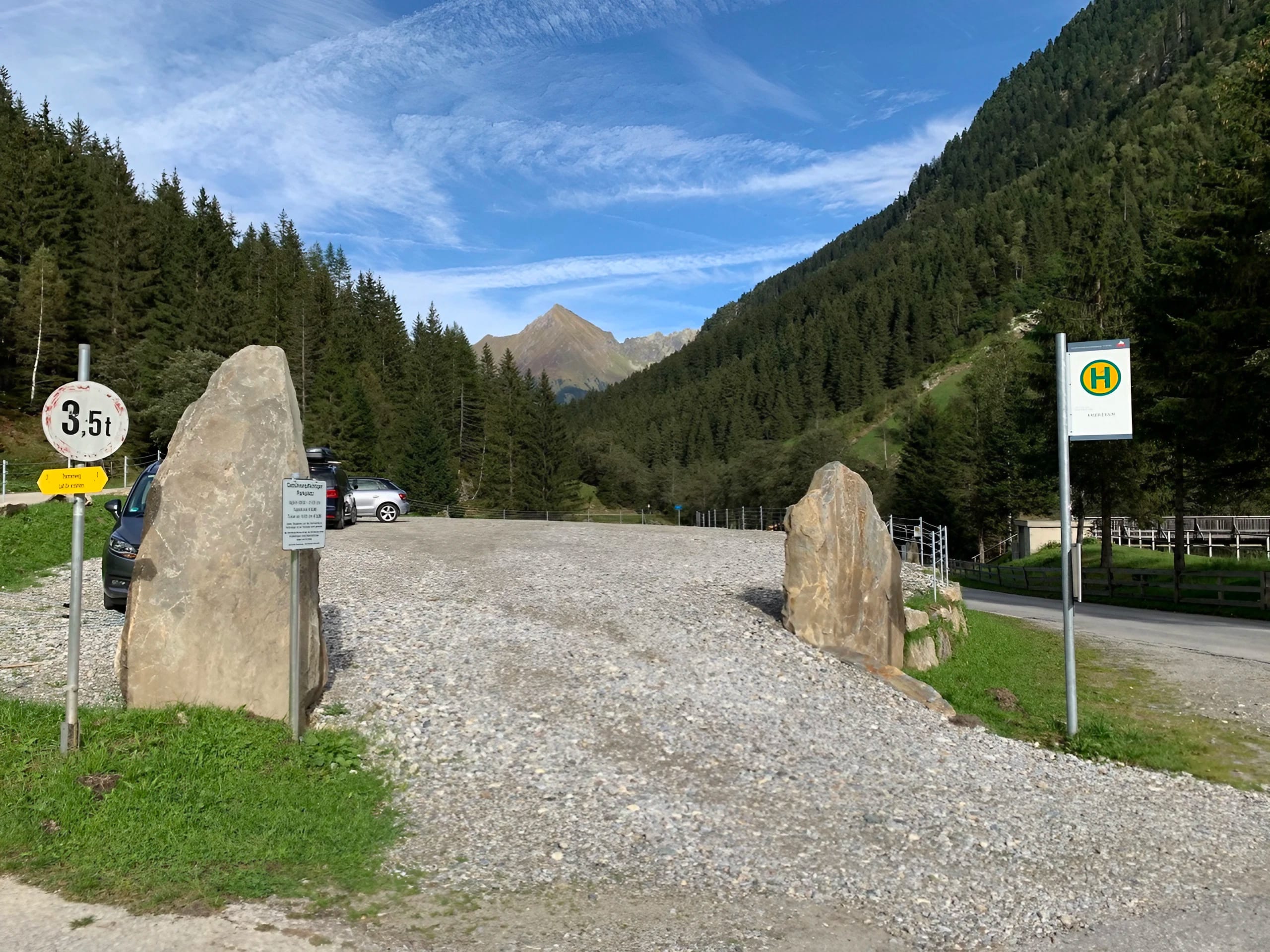 Berglandschaft mit bewaldeten Hügeln, blauer Himmel und Wolken, Parkplatz mit Autos und Schildern im Vordergrund.
