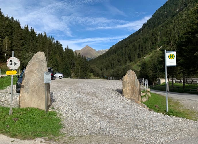 Mountain landscape with forested hills, blue sky, clouds, and a parking lot with cars and signs in front.