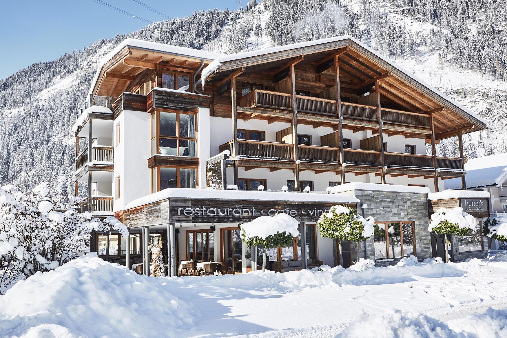 Mehrstöckiges Chalet im Schnee mit Holz und weißen Wänden, Restaurant- und Café-Schilder, umgeben von verschneiter Landschaft.