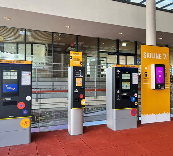 Automaten für Skipass-Tickets vor einem Gebäude mit Glasfassade und roten Bodenfliesen.