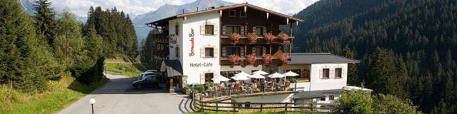 The Hotel Bergkristall is seen from the outside, nestled within a wide meadow, surrounded by forest and mountains. The sky is now cloudy, giving the scene a calm and subdued atmosphere