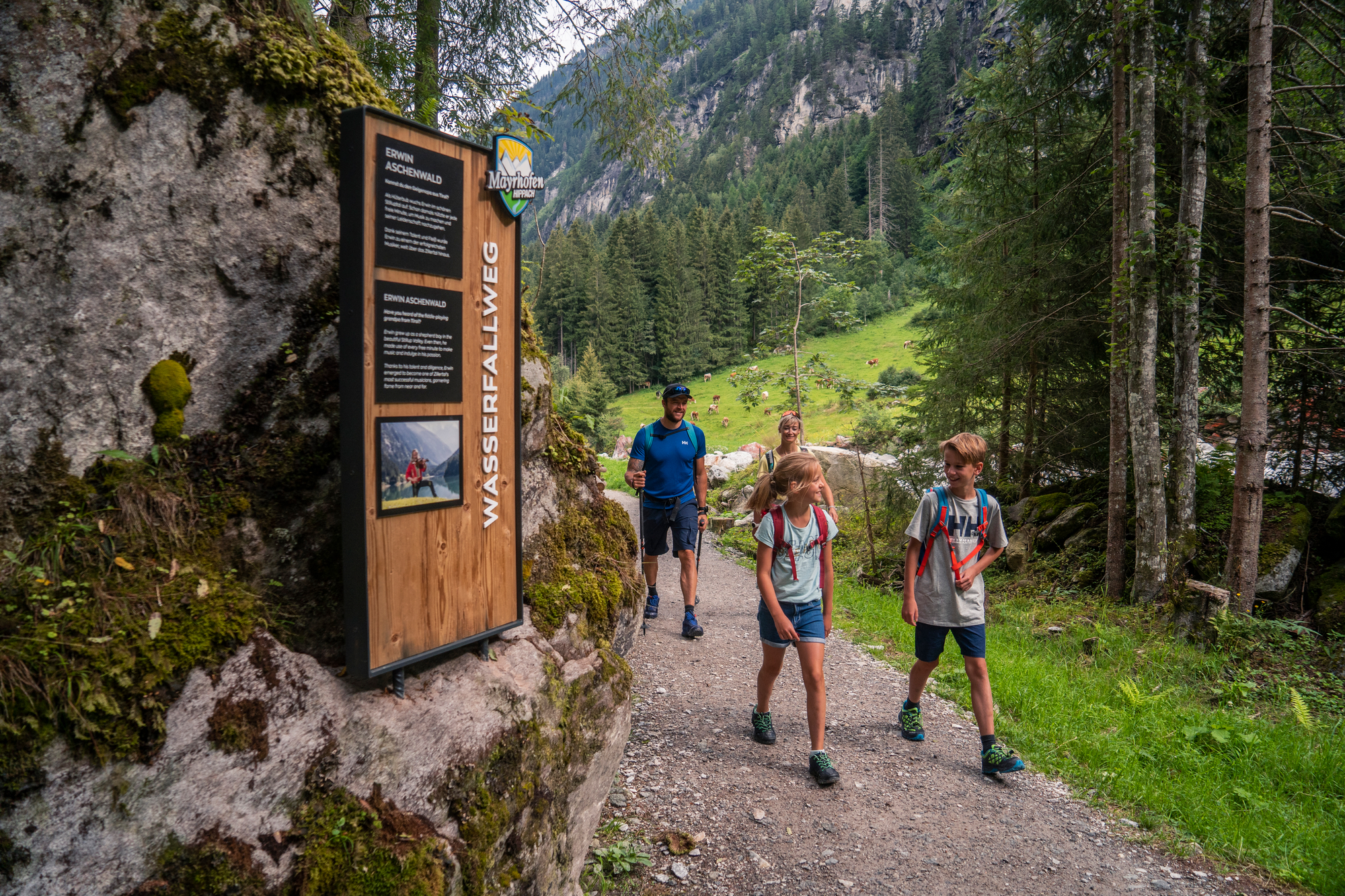 Familie wandert am Wasserfallweg im Stilluptal bei Mayrhofen. Der Weg führt durch einen Waldabschnitt, vorbei an einem hölzernen Infopoint am Felsen. Im Hintergrund öffnet sich der Blick auf grüne Almen und steile Berghänge.