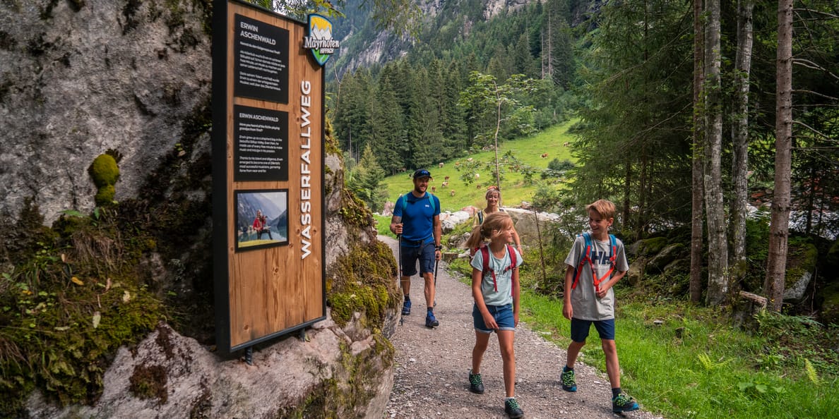 Stilluptal Waterfall Trail Family hiking along the Waterfall Trail in the Stilluptal valley near Mayrhofen. The path leads through a forest past a wooden info board on a rock. In the background, green alpine pastures and steep mountain slopes come into view