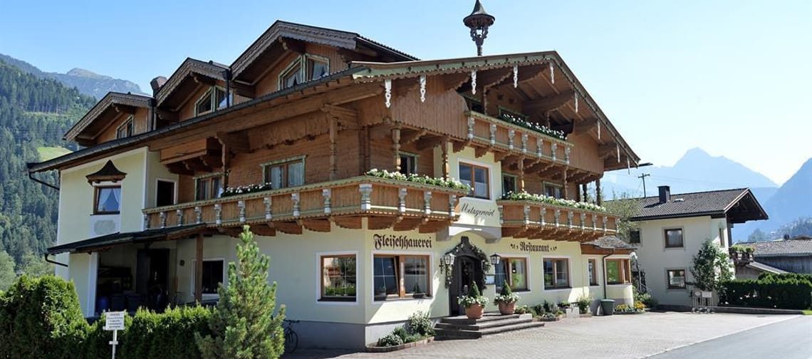 Das Bild zeigt den Metzgerwirt von außen. Im Hintergrund ragen eindrucksvolle Berge in den blauen Himmel, was der Szene eine typische alpine Kulisse verleiht.