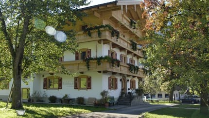 Ein mehrstöckiges Haus im alpinen Stil mit Balkon, umgeben von Bäumen und Garten, bei Sonnenlicht.