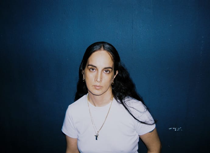 Young woman with dark hair against a blue wall, wearing a white T-shirt and necklace, looking at the camera.