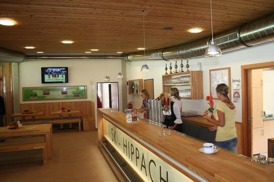 Cozy interior of the clubhouse at Lindenstadion Hippach with wooden ceiling, long counter, seating areas, and guests; home of SK Raika Hippach in Tyrol.