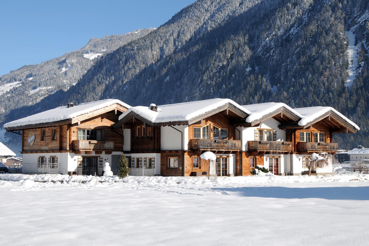 A snowy chalet in front of a forested mountain, with wooden facades and snow on the roof.