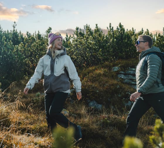 Zwei Wanderer in Outdoor-Bekleidung auf einem Weg in einer bewaldeten Berglandschaft bei Sonnenuntergang.