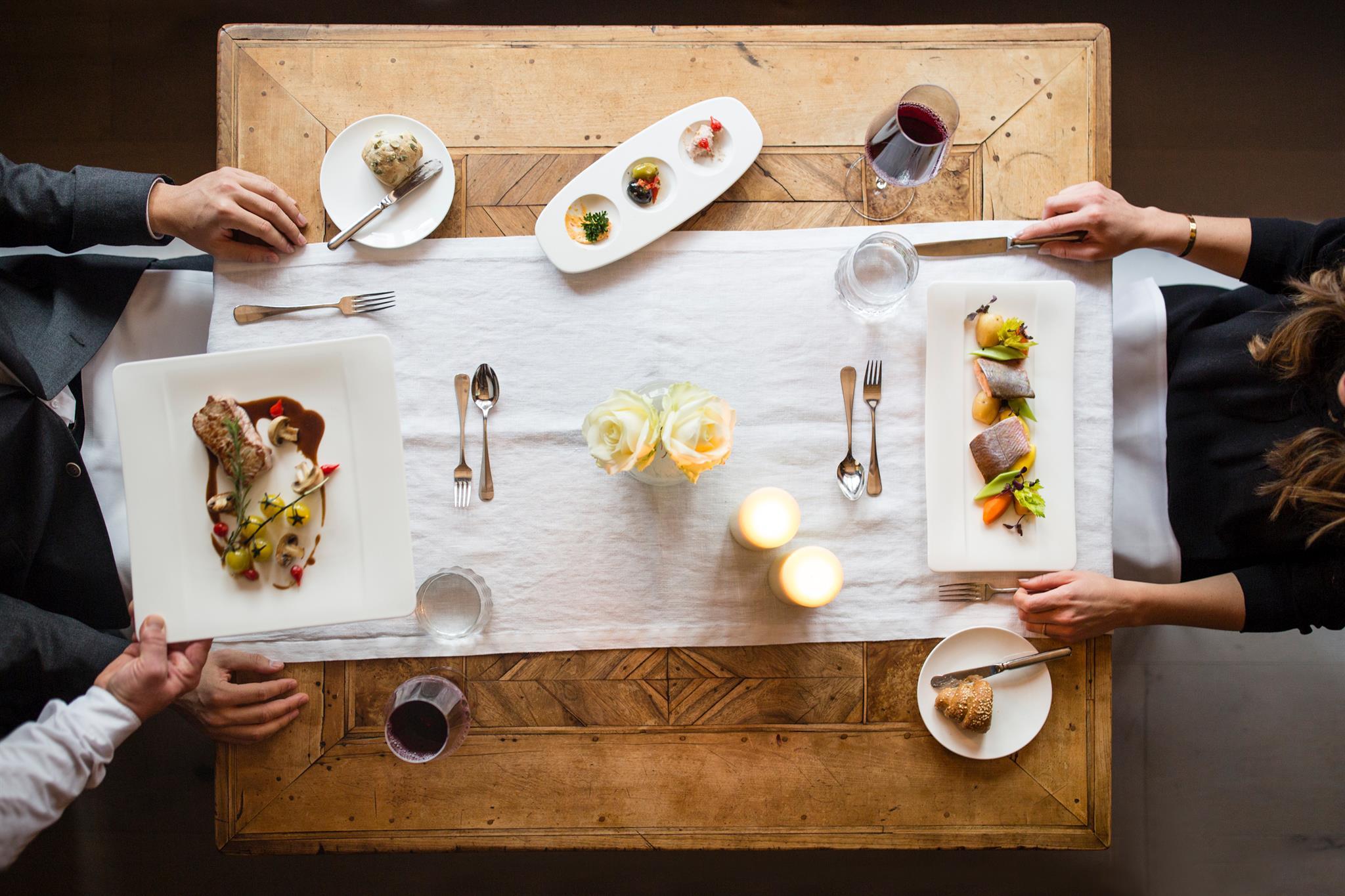 Two people dining at a wooden table with plates, wine, and candles, viewed from above.