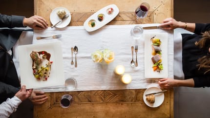 Two people dining at a wooden table with plates, wine, and candles, viewed from above.