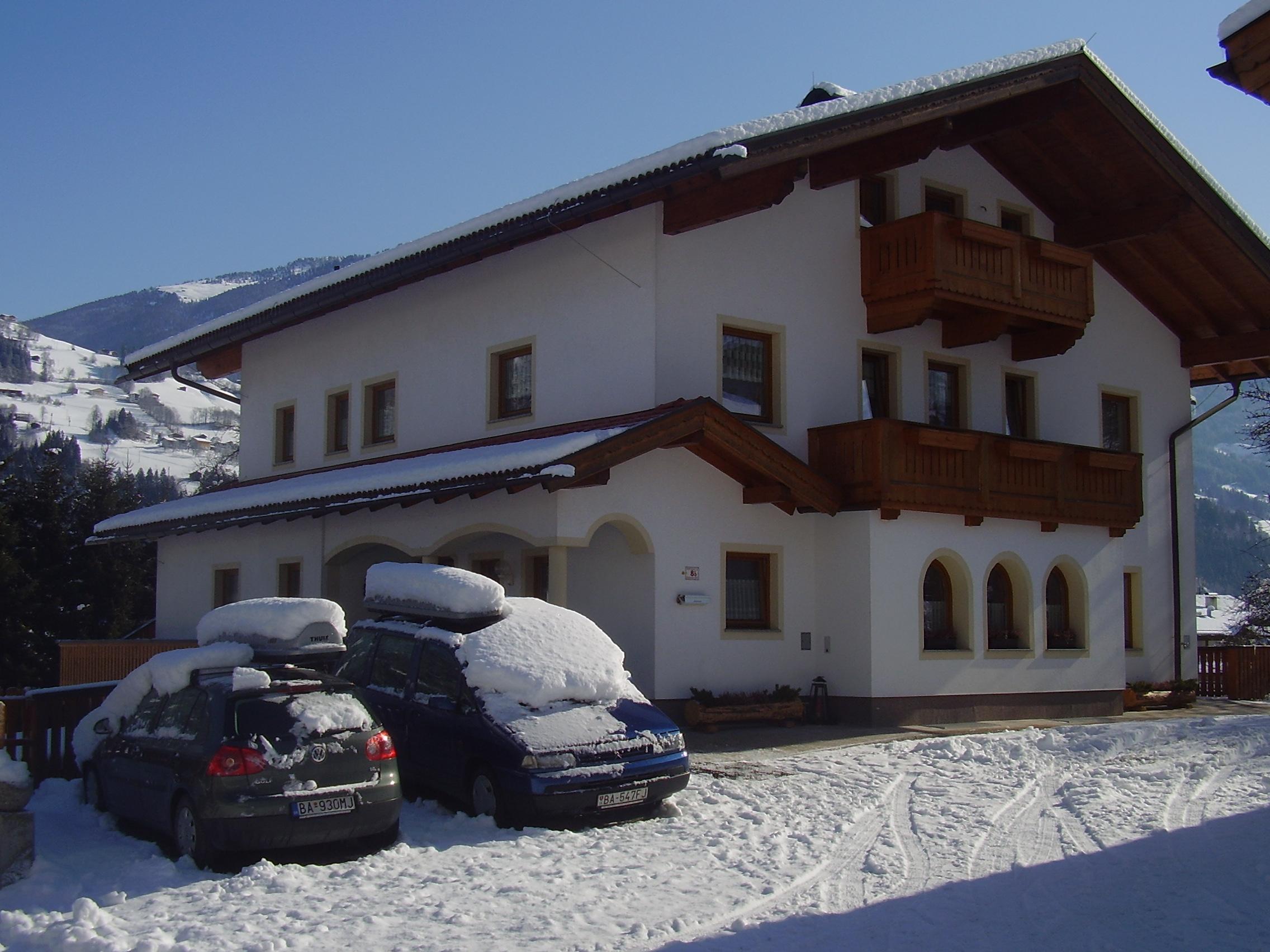 A multi-story house with white walls, wooden balconies, and snow-covered roof in a mountain winter scene.