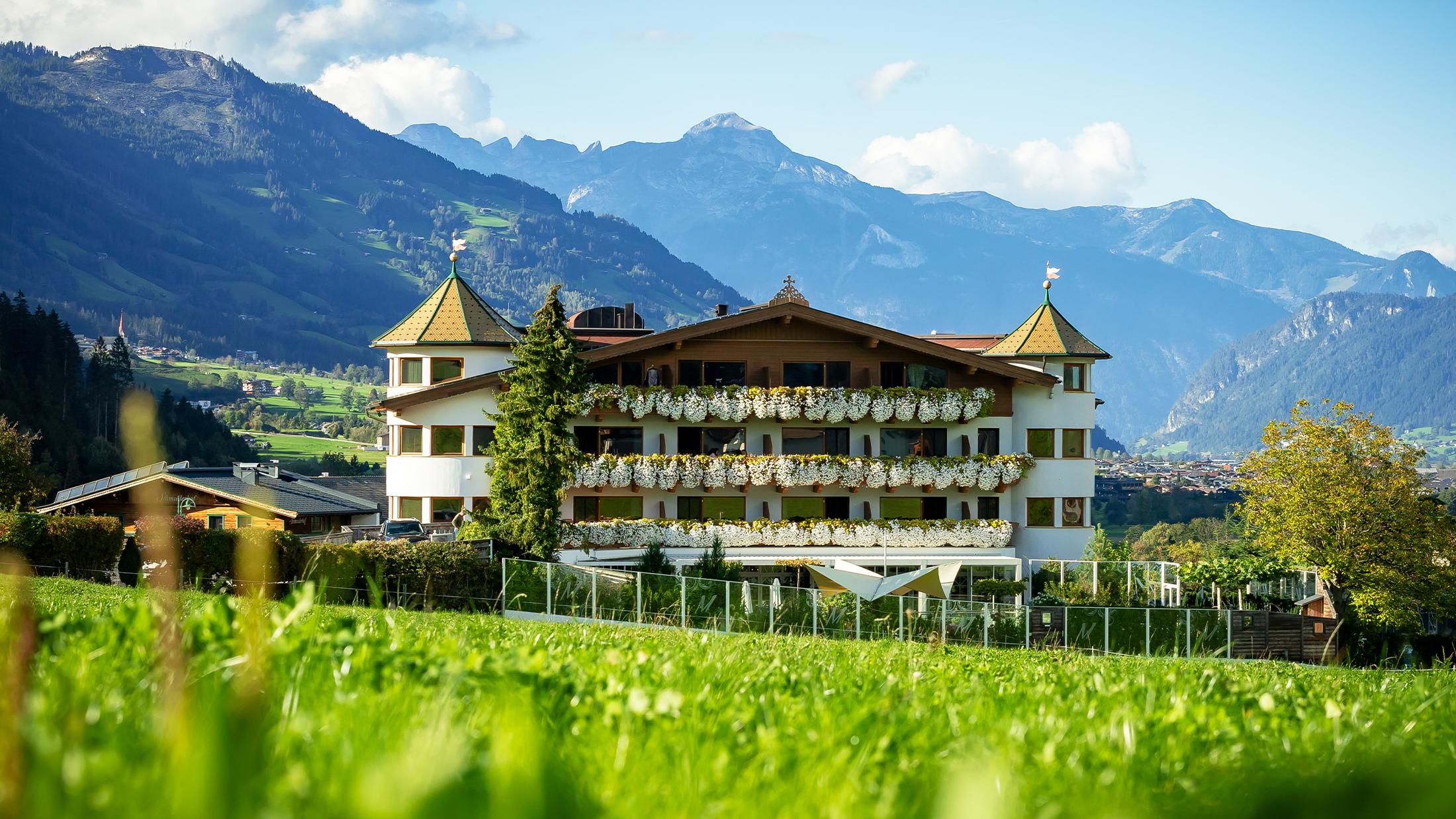 Ein Haus mit Blumen auf Balkonen vor einer Bergkulisse in den Alpen, bei sonnigem Himmel.