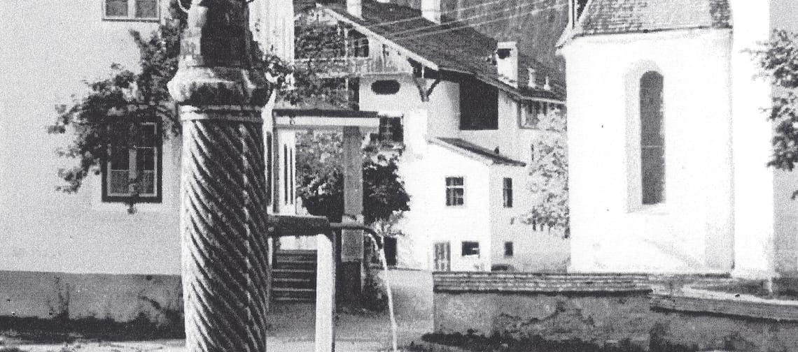 Schwarzweißfoto von 1928: Marienbrunnen in Mayrhofen – Brunnen mit Säule und Becken im Vordergrund; dahinter Pfarrkirche und Widum, weitere Häuser und Bergflanke.