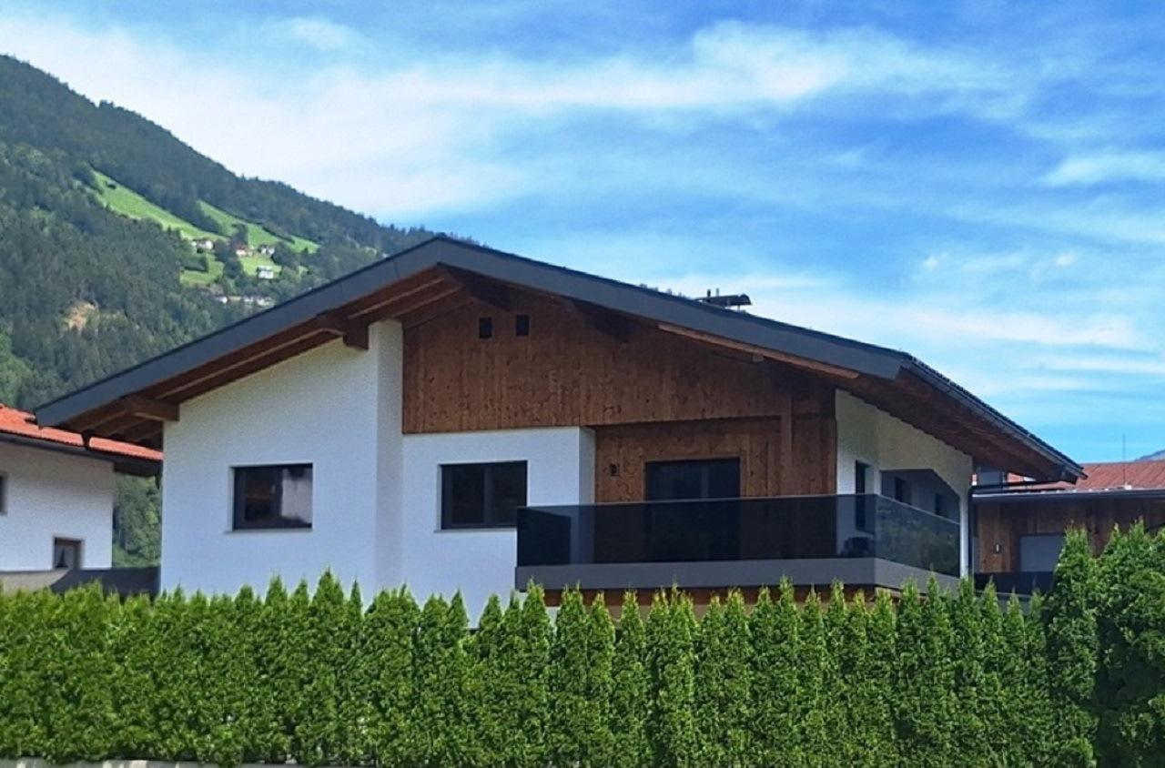 Modern house with white walls, wooden accents, and balcony, against green hills and blue sky.