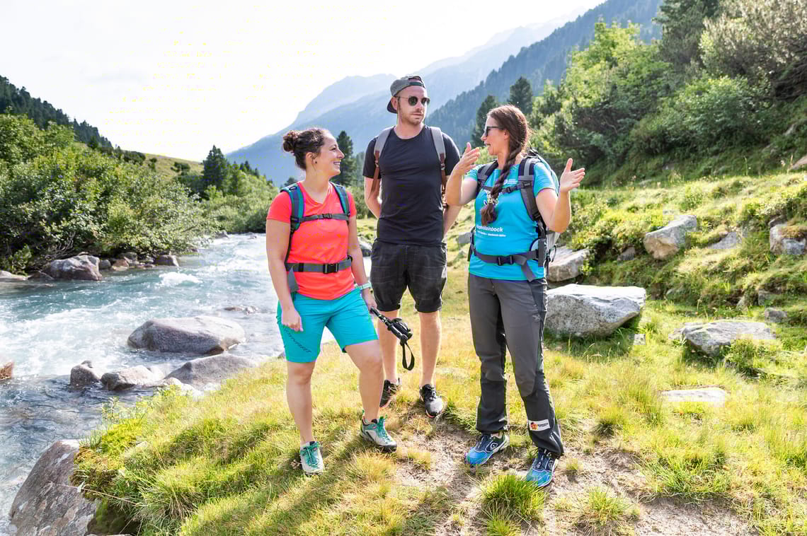 Geführte Wanderung am Schlegeisspeicher Drei Wanderer mit Rucksäcken gehen auf einem Pfad im Naturpark Zillertaler Alpen im Schlegeis. Umgeben von grünen Bäumen und einem Fluss genießen sie die sonnige Landschaft.