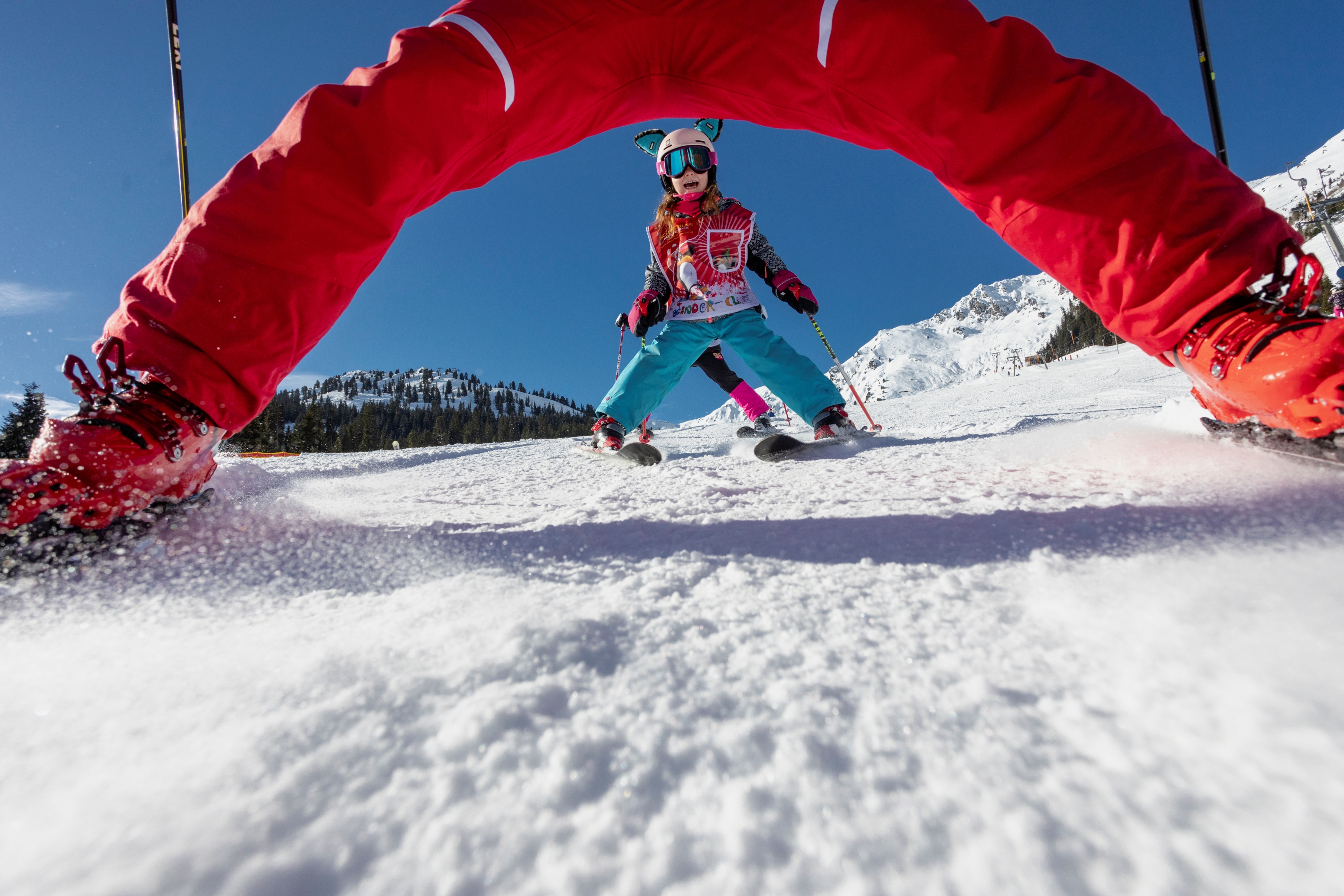 Ein Kind beim Skikurs, trägt Helm und Skibrille, steht auf Skiern im Schnee, umgeben von Bergen und blauer Himmel.