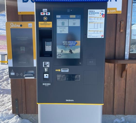 Automatischer Ticketautomat vor einer Holzwand, mit Bildschirm, Kartenleser und Schneefall im Hintergrund.