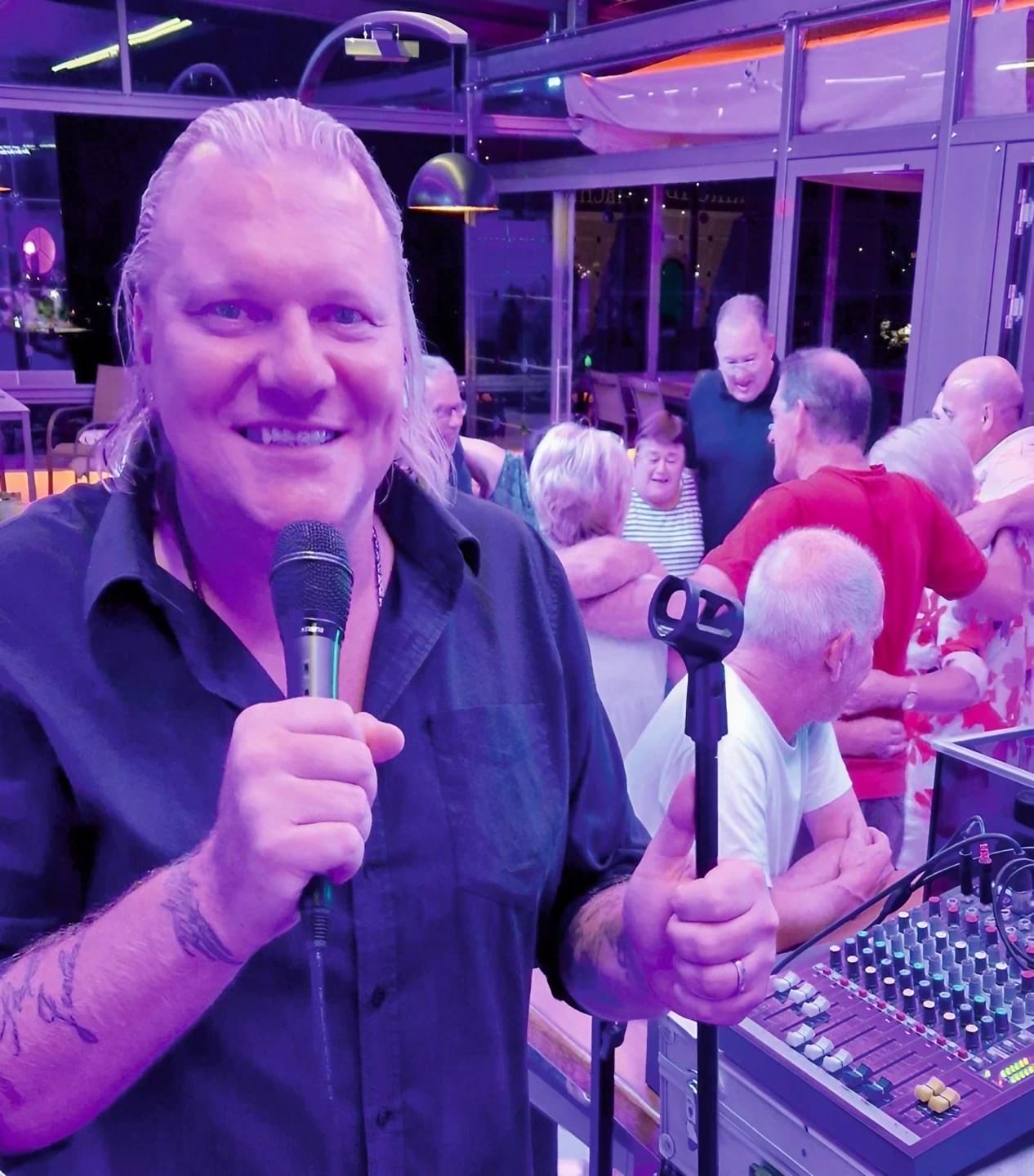 Das Bild zeigt einen DJ mit Mikrofon und Gästen im Hintergrund in einem Lokal