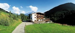 The Hotel Bergkristall is seen from the outside, nestled within a wide meadow, surrounded by forest and mountains. The sky is now cloudy, giving the scene a calm and subdued atmosphere