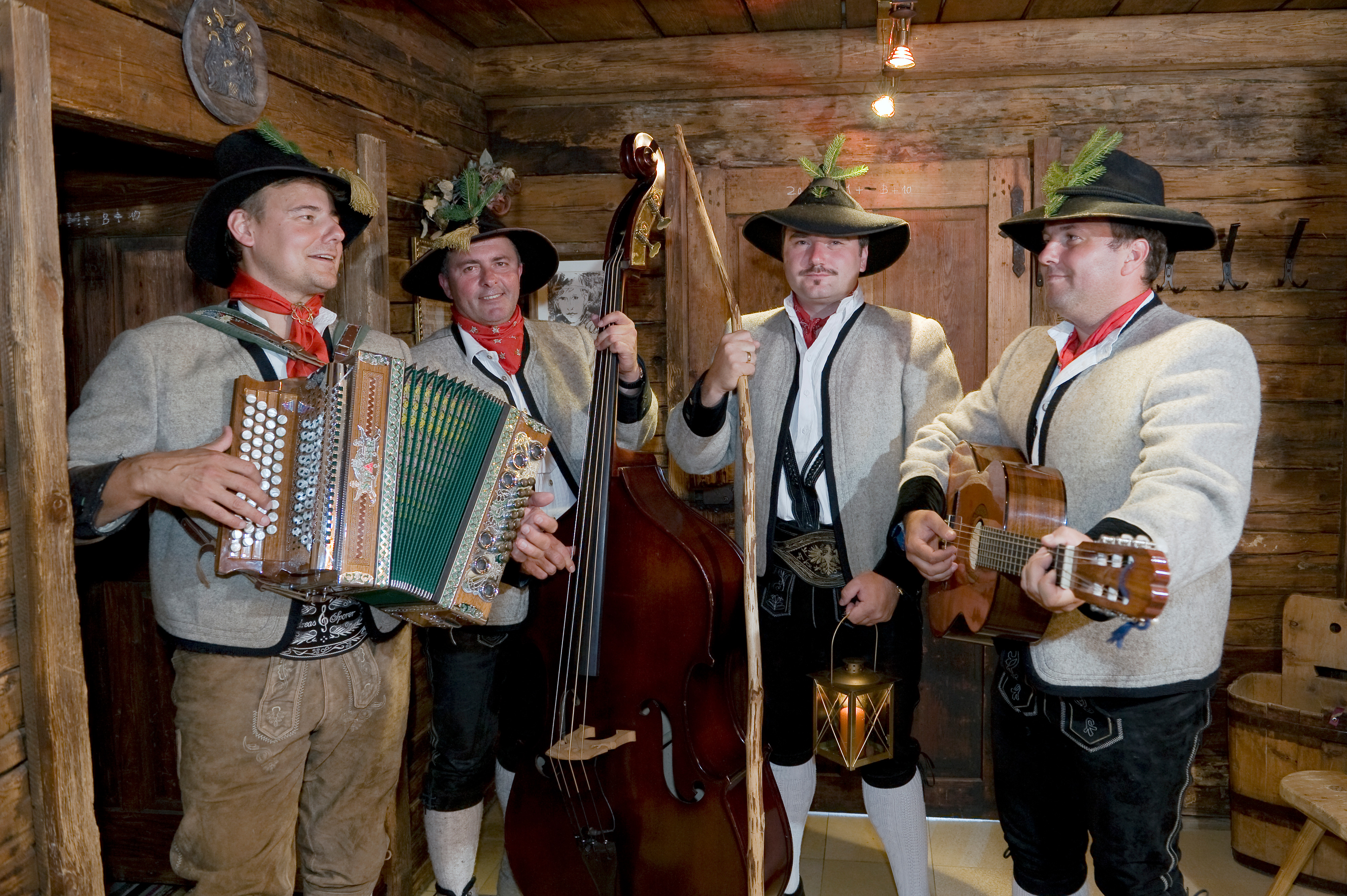 A traditional folk band plays music. The men wear hats, shirts, and leather trousers. They play accordion, guitar, and double bass in a wooden room. Everyone enjoys the music.