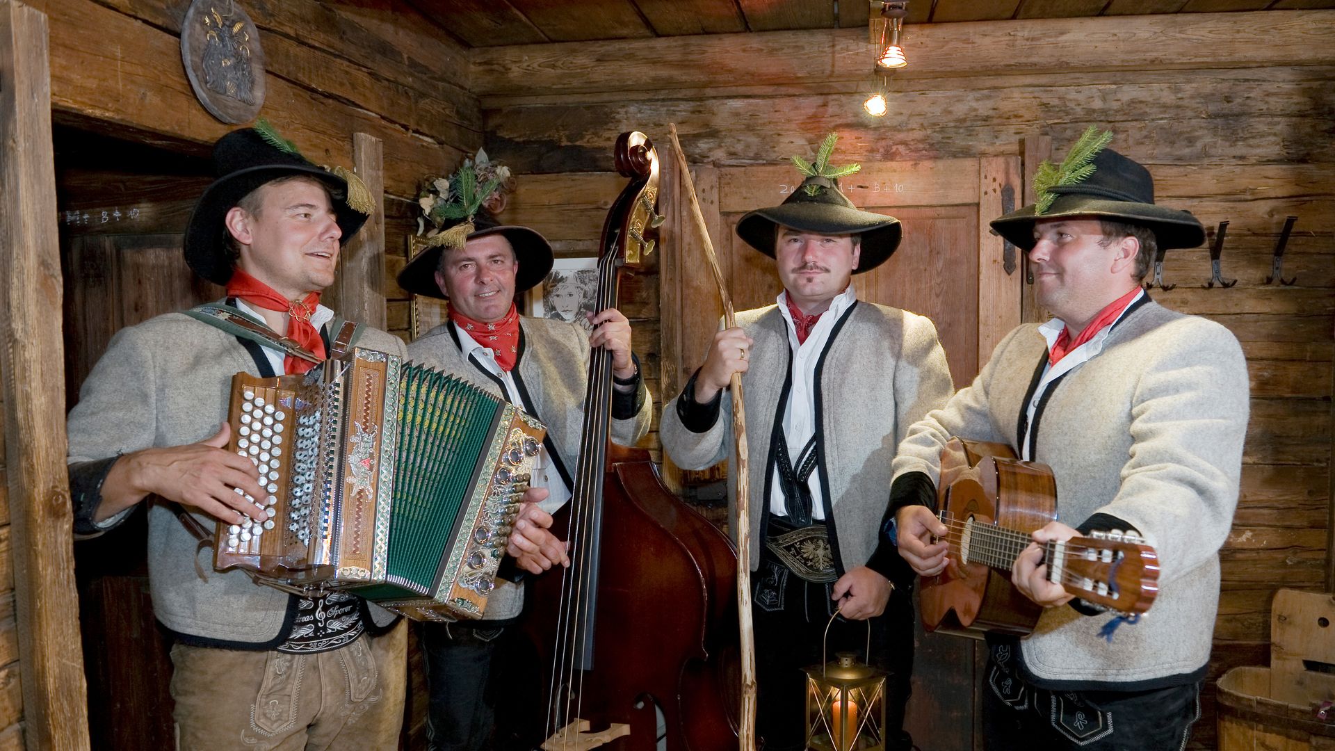 A traditional folk band plays music. The men wear hats, shirts, and leather trousers. They play accordion, guitar, and double bass in a wooden room. Everyone enjoys the music.