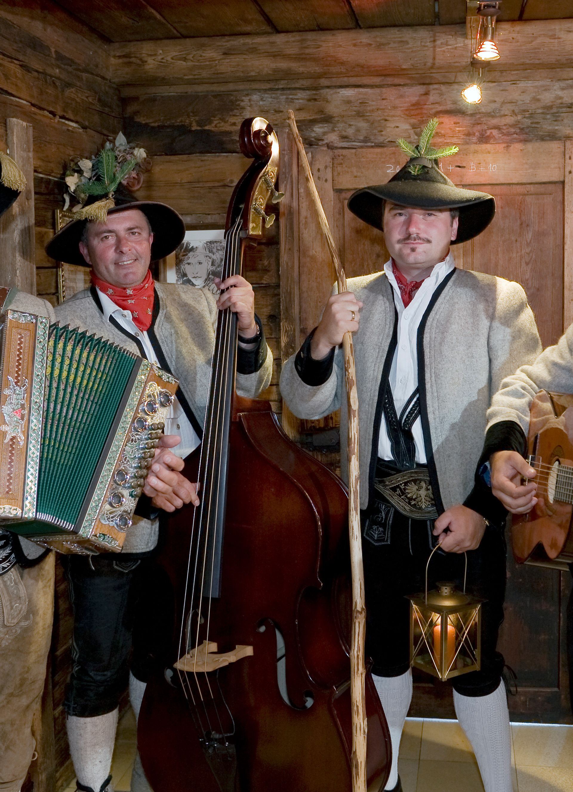 A traditional folk band plays music. The men wear hats, shirts, and leather trousers. They play accordion, guitar, and double bass in a wooden room. Everyone enjoys the music.