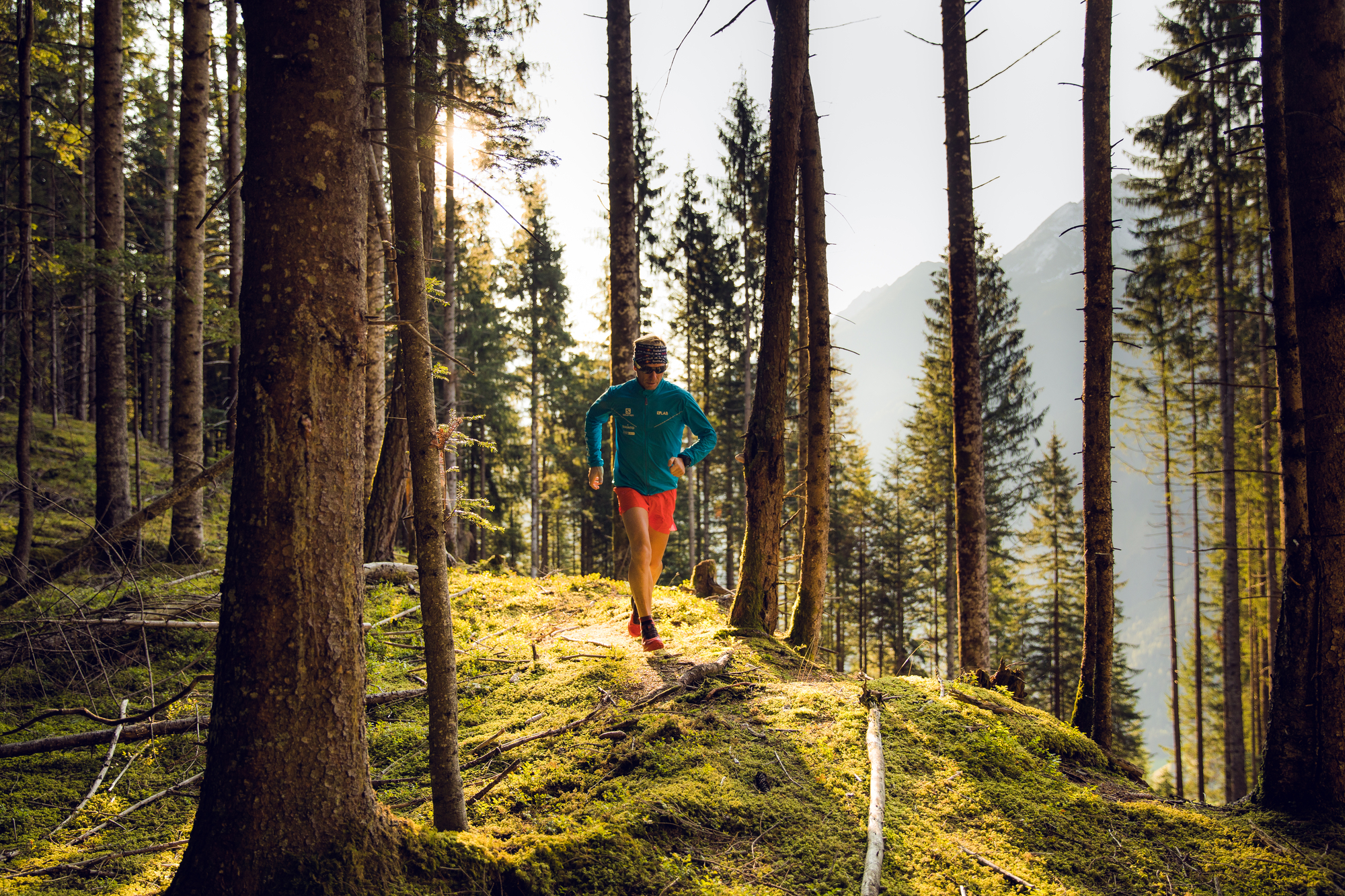 Eine Person beim Trailrunning auf einem weichen Waldpfad in der Ferienregion Mayrhofen-Hippach, Zillertal. Sonnenlicht fällt durch die hohen Bäume, im Hintergrund sind Berge zu sehen. Naturerlebnis und sportliche Herausforderung.