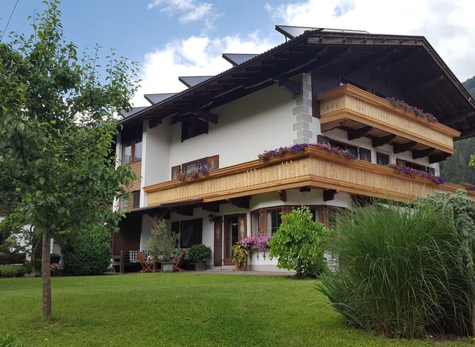 Ein mehrstöckiges Haus mit Holzbalconnen, Blumen und umgeben von Bäumen und grüner Wiese in den Bergen.
