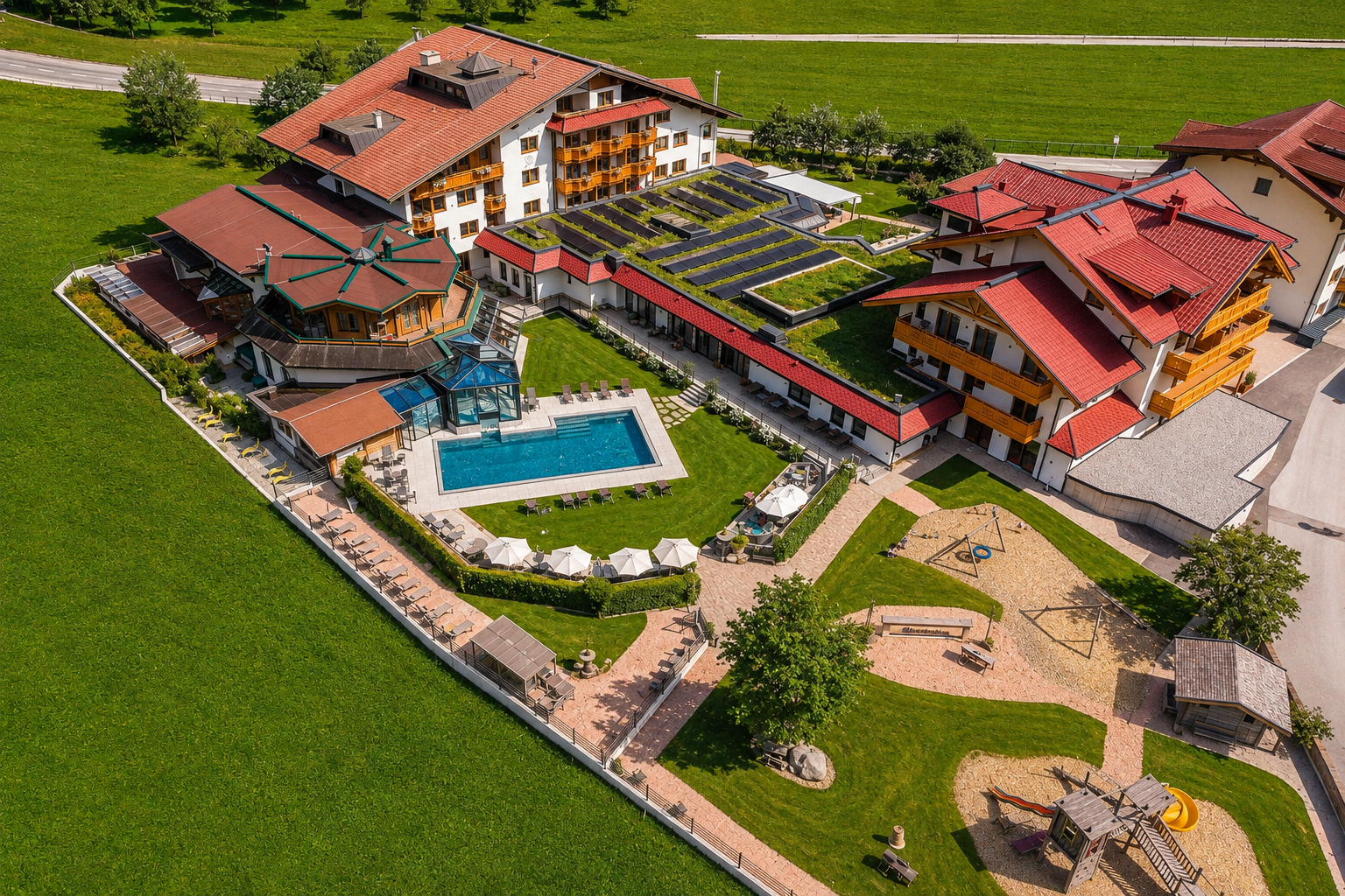 Luftaufnahme eines Hotels mit Pool, Spielplatz und grünen Gärten, umgeben von Wiesen und Bäumen.