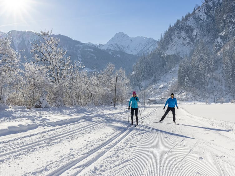 Two cross-country skiers on the Burgstall trail in Mayrhofen-Hippach, surrounded by snowy trees and mountain scenery in bright sunshine.